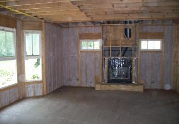 Empty unfinished room with wood-paneled walls, exposed ceiling beams, windows, and a fireplace opening.