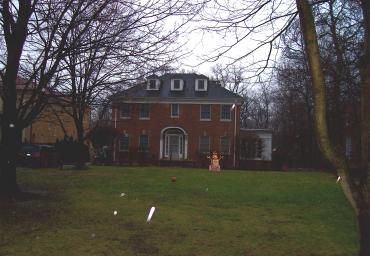Large brick house with white trim on a grassy lawn, framed by bare trees on a cloudy day