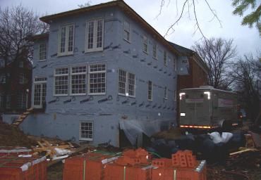 Damaged two-story house with a bus or truck beside it and orange safety fencing in front