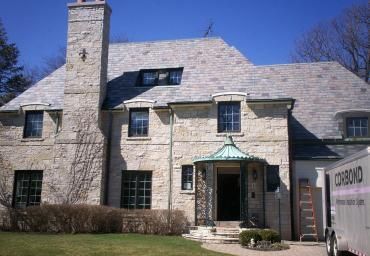 Stone house with a chimney, green-trimmed windows, and a small entrance gate under a blue sky