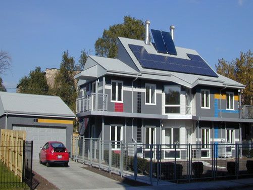 Modern gray house with solar panels, a red car, and a fenced yard under a clear blue sky