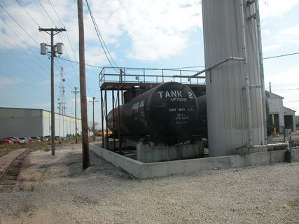Black rail tank car beside industrial silos and utility poles in a gravel yard