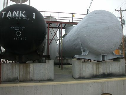 Two industrial storage tanks on concrete bases, one black labeled “TANK 1” and one covered in gray paint.