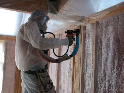 Worker in protective suit spray-insulating a wall inside a room under construction.