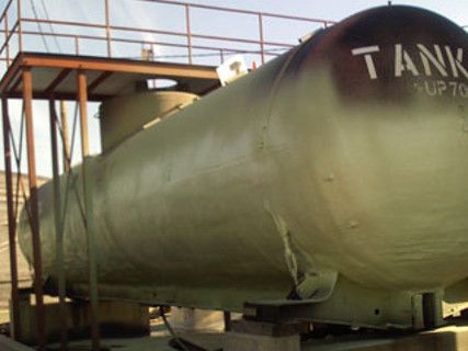 Large olive-green tank labeled “TANK” on a metal platform outdoors, with stairs and railings nearby