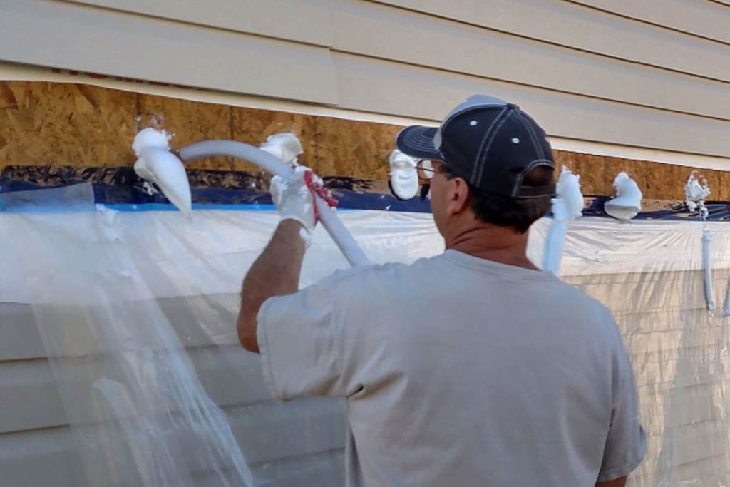 Man spraying blue paint on house siding with a sprayer while covering windows with plastic sheets
