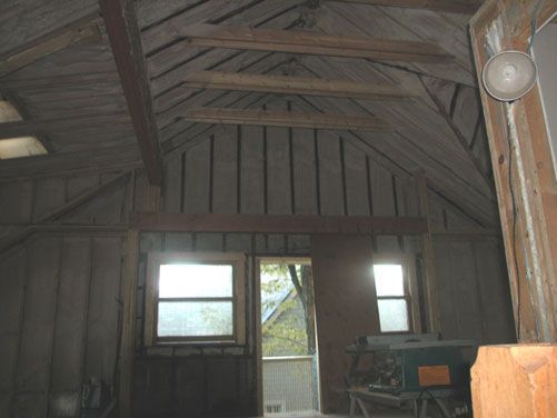 Rustic wooden shed interior with exposed rafters, small windows, and a door opening to greenery