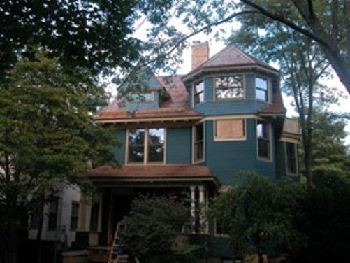 Two-story teal Victorian house with red roof and a covered porch, surrounded by trees