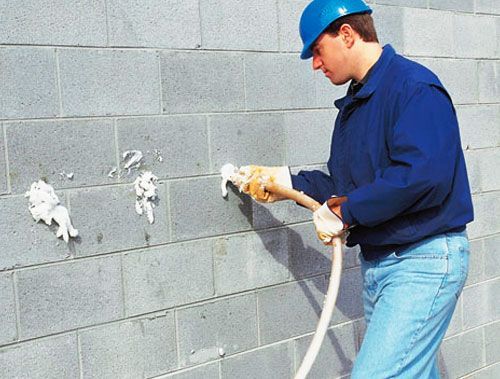 Worker in blue uniform and hard hat spraying insulation onto a gray cinderblock wall.
