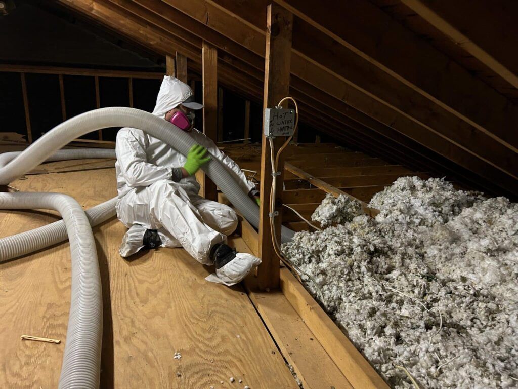 Worker in white protective suit insulating an attic, surrounded by ducts and loose insulation
