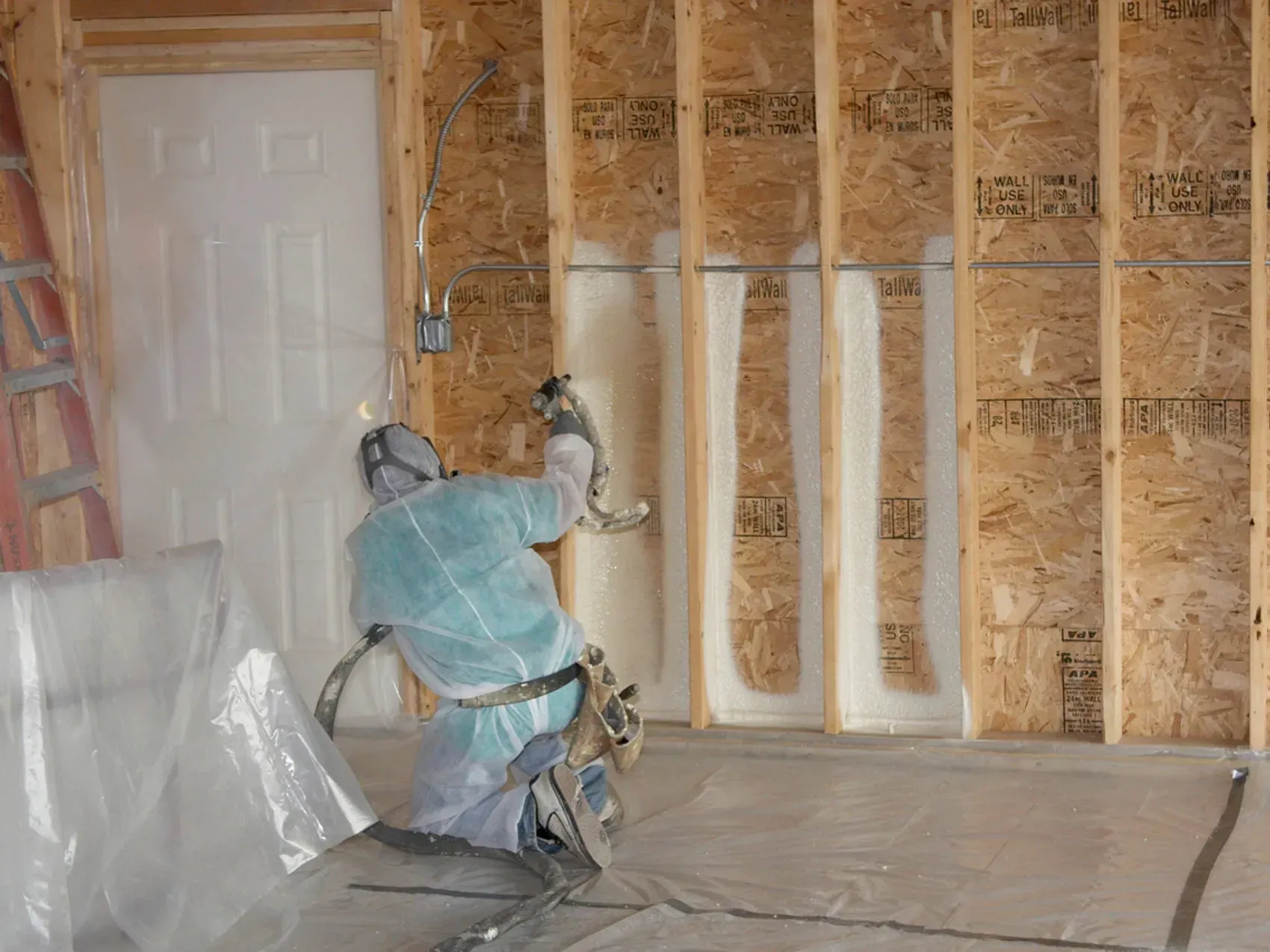 Worker spraying insulation foam inside an unfinished room with exposed wall studs.