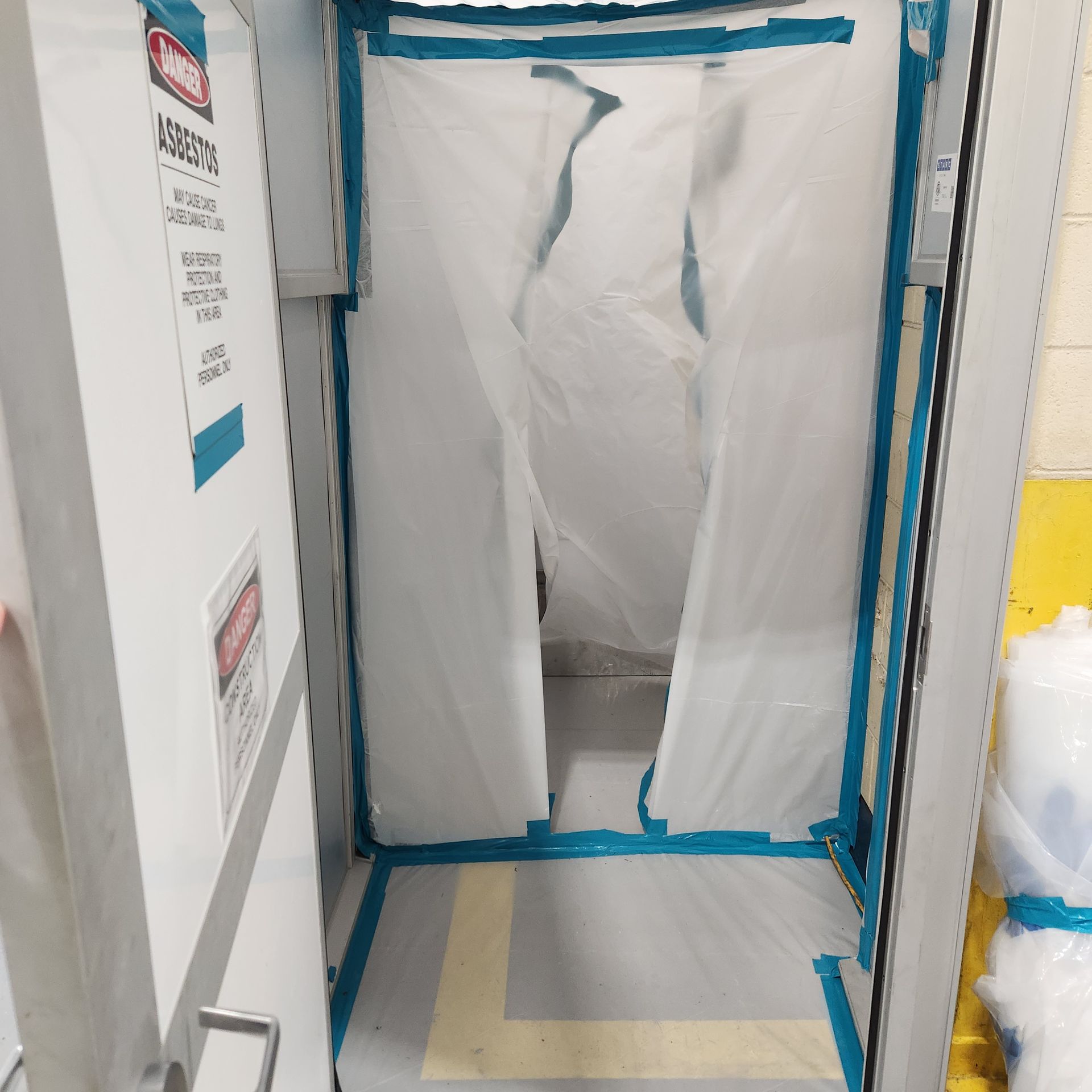 A doorway sealed with plastic sheeting and blue tape. A warning sign is visible on the door.