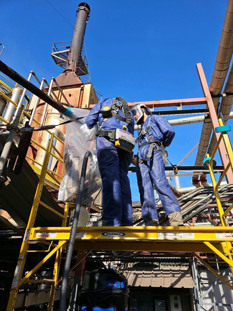 Two workers in blue coveralls on a scaffold at an industrial site, inspecting equipment.