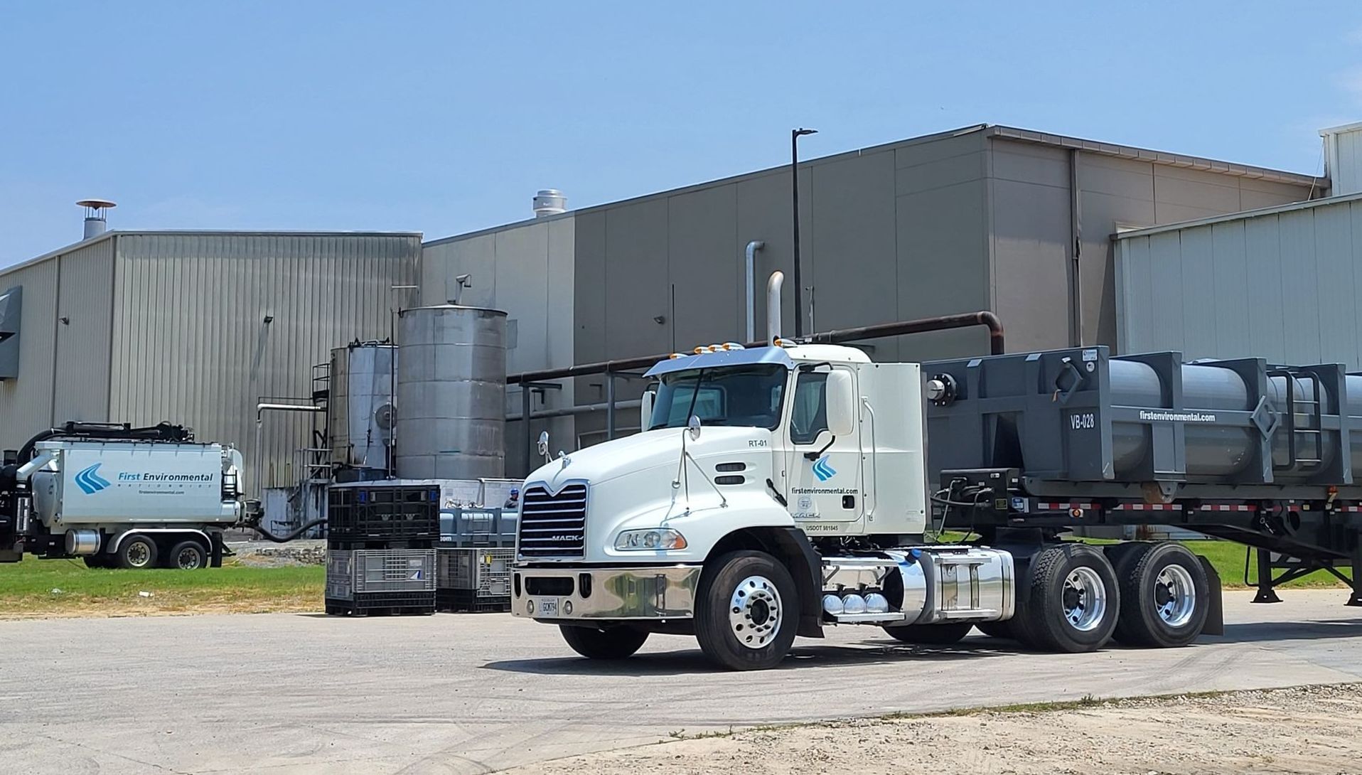 White semi-truck with tanker trailer in front of a factory building under a bright sky.