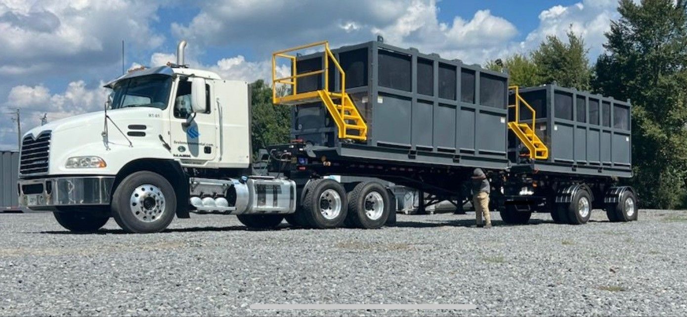 White Mack truck towing two large, gray trash containers with yellow access stairs, parked outside.