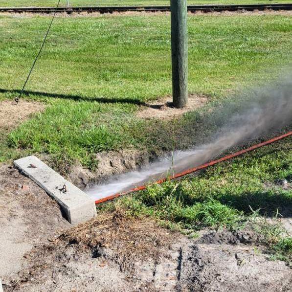 A pressurized hose sprays water into a ditch, near a wooden pole and green grass.