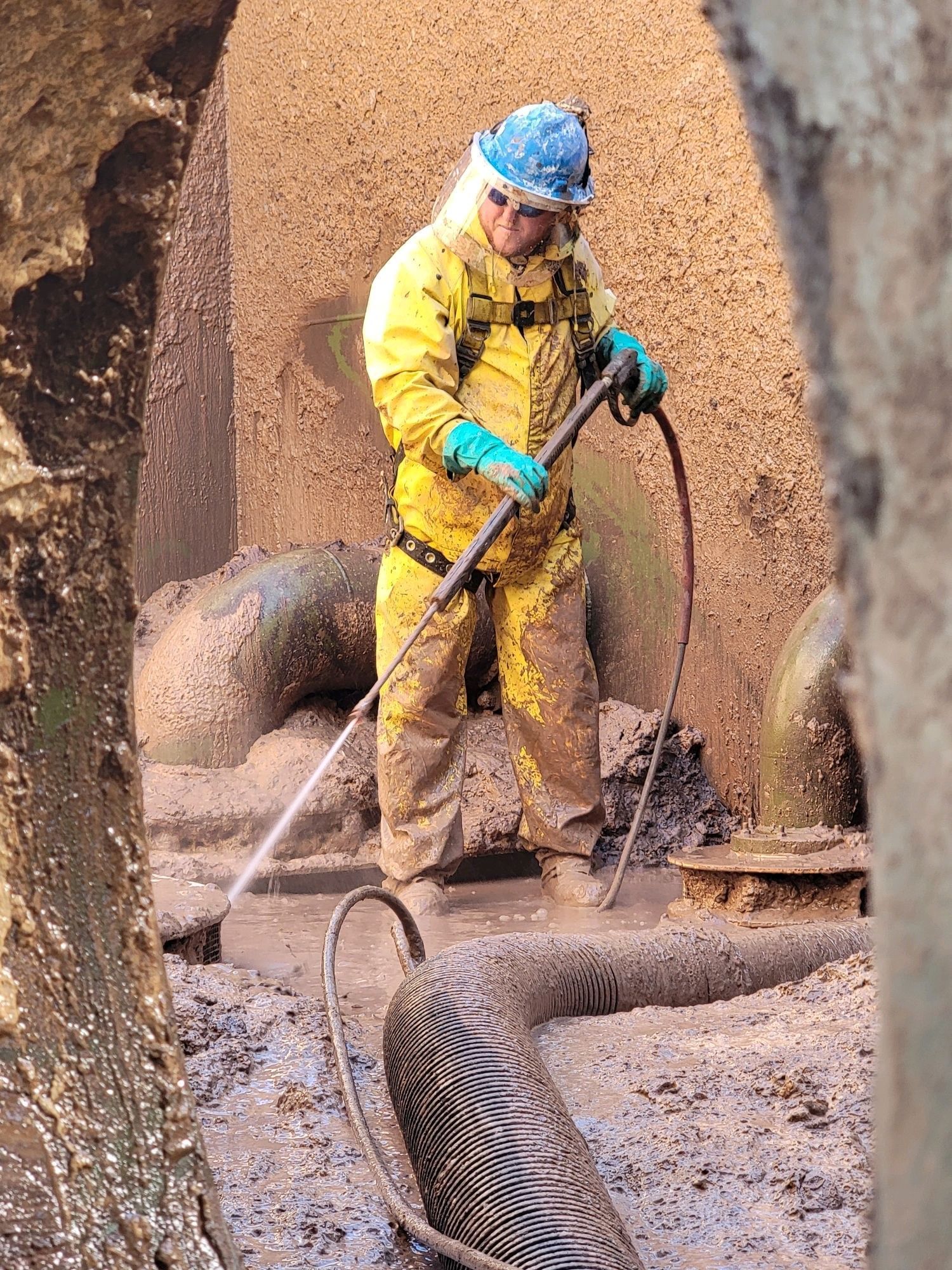 Person in yellow hazmat suit pressure-washing muddy pipes in a confined space.