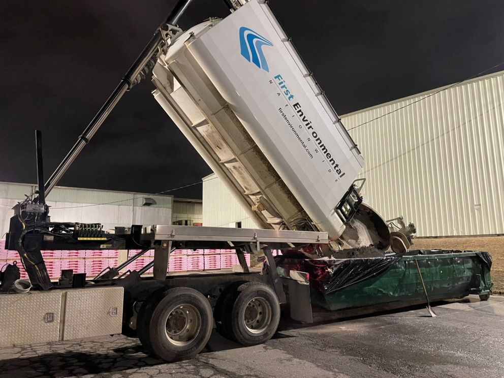 A First Environmental truck dumping waste into a green dumpster at night.