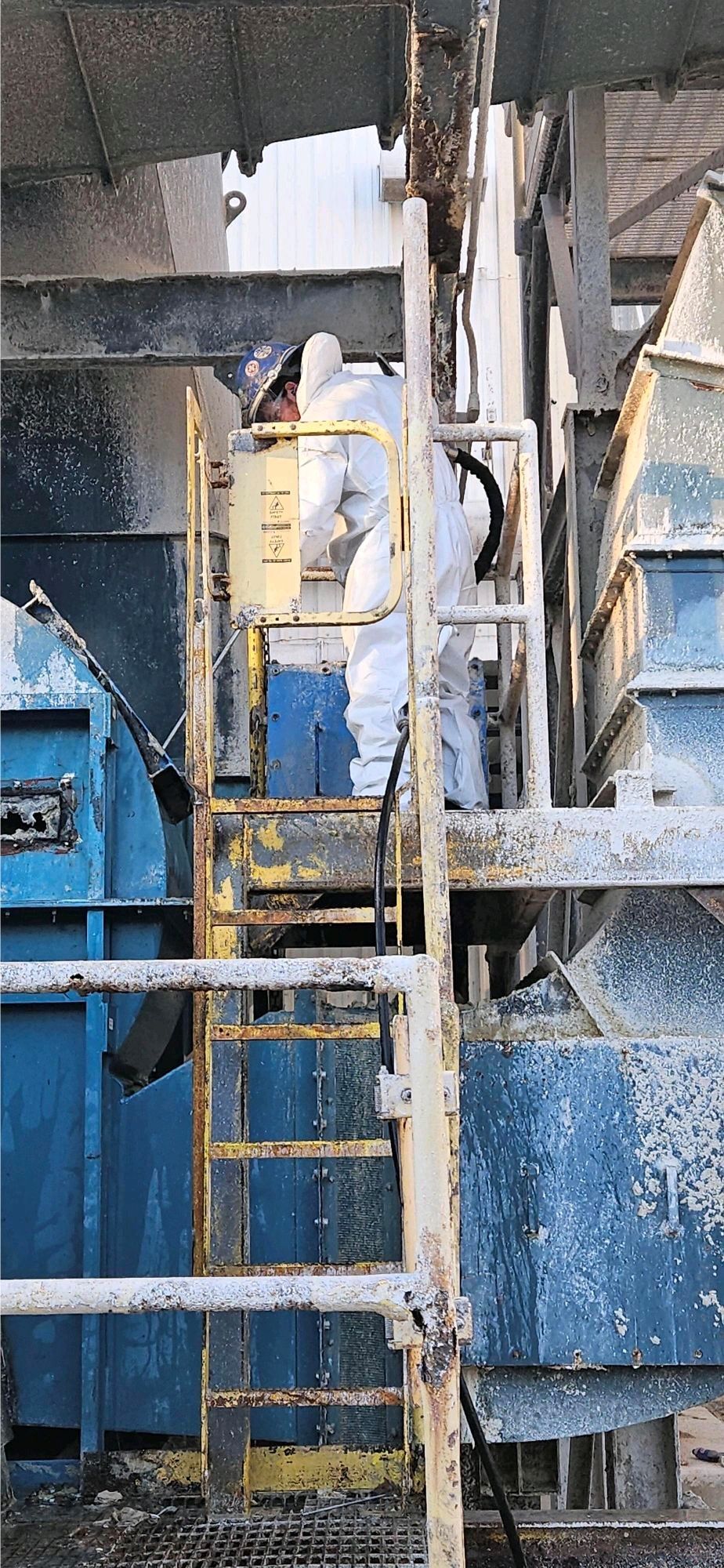 A First Environmental employee in a protective suit is standing on a ladder in a factory.