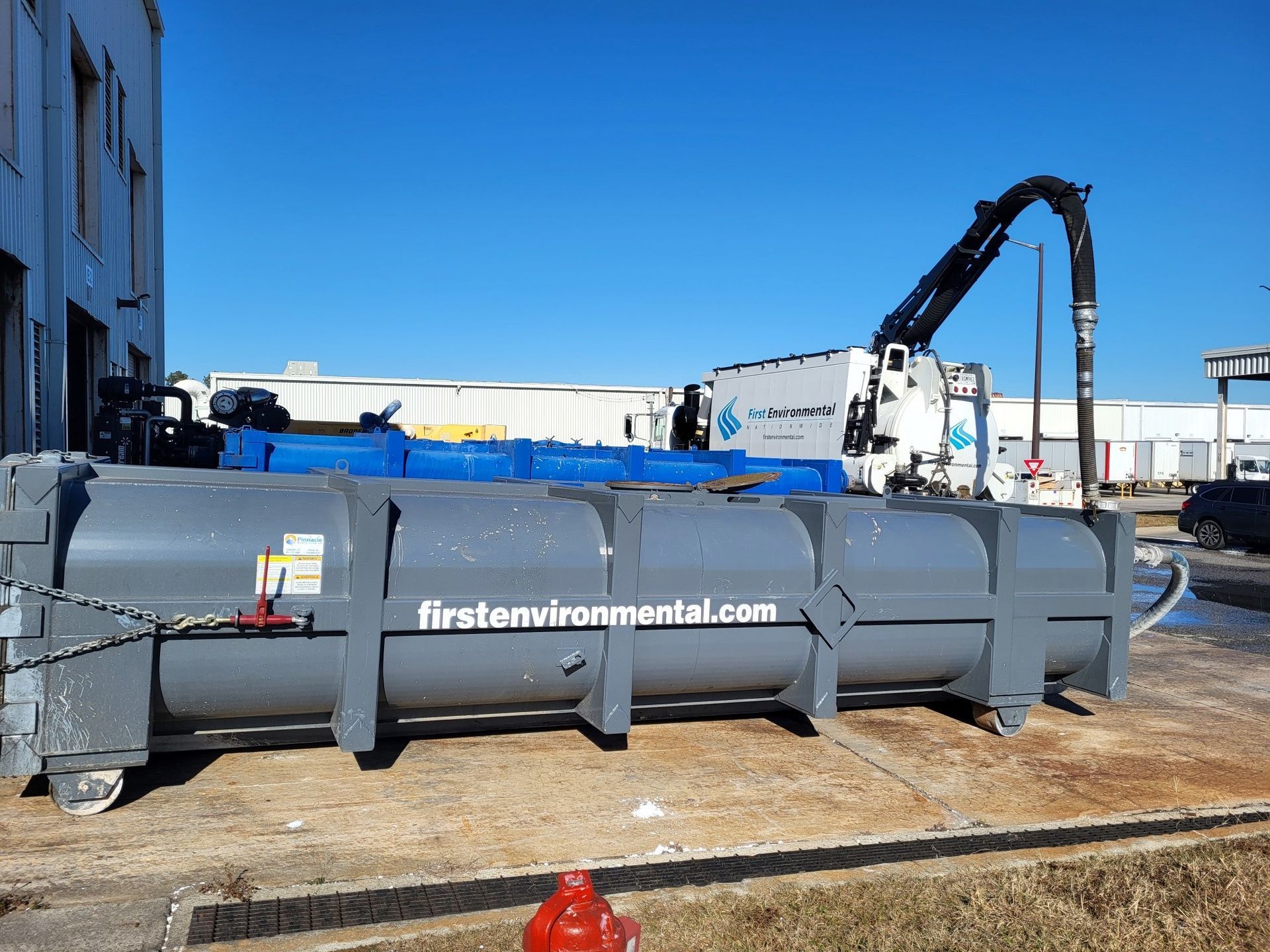 A large gray container with the website firstenvironmental.com being loaded by a crane.