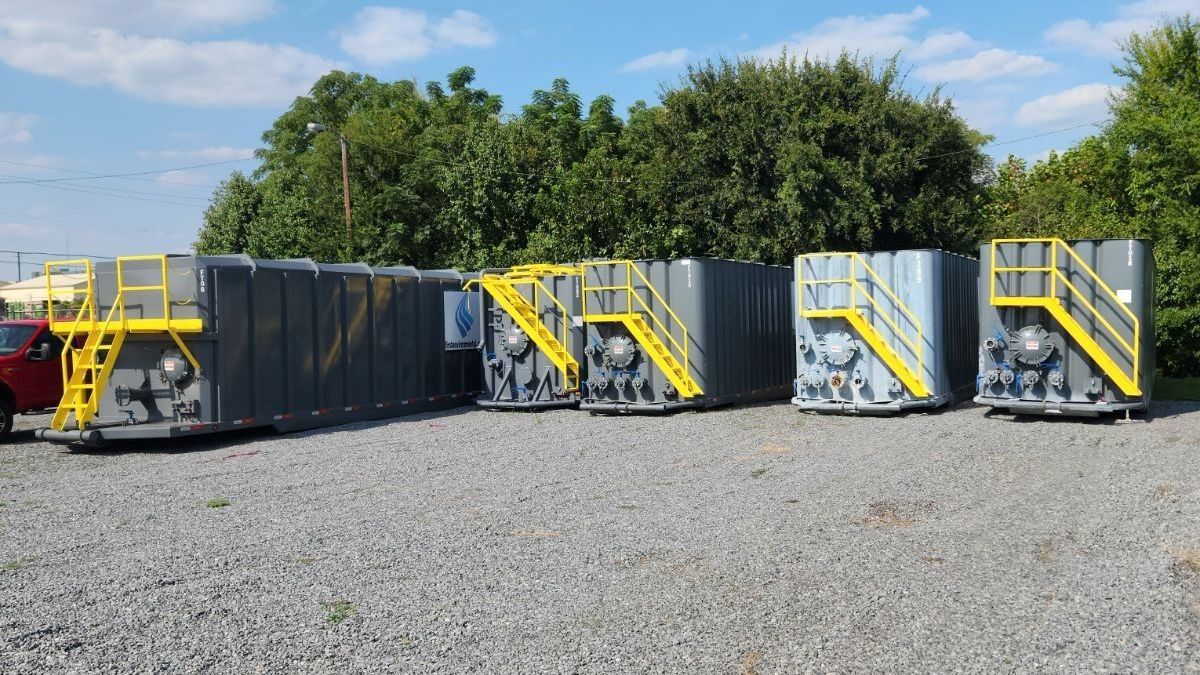 Five dark gray industrial tanks with yellow stairs, parked on gravel under a blue sky.