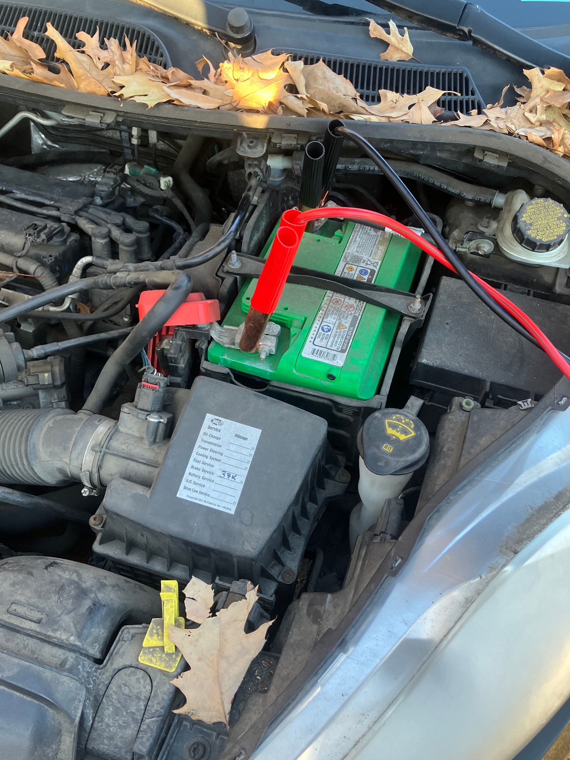 Car engine bay with a green battery and red cable, debris on the hood, and a small fire near the windshield.
