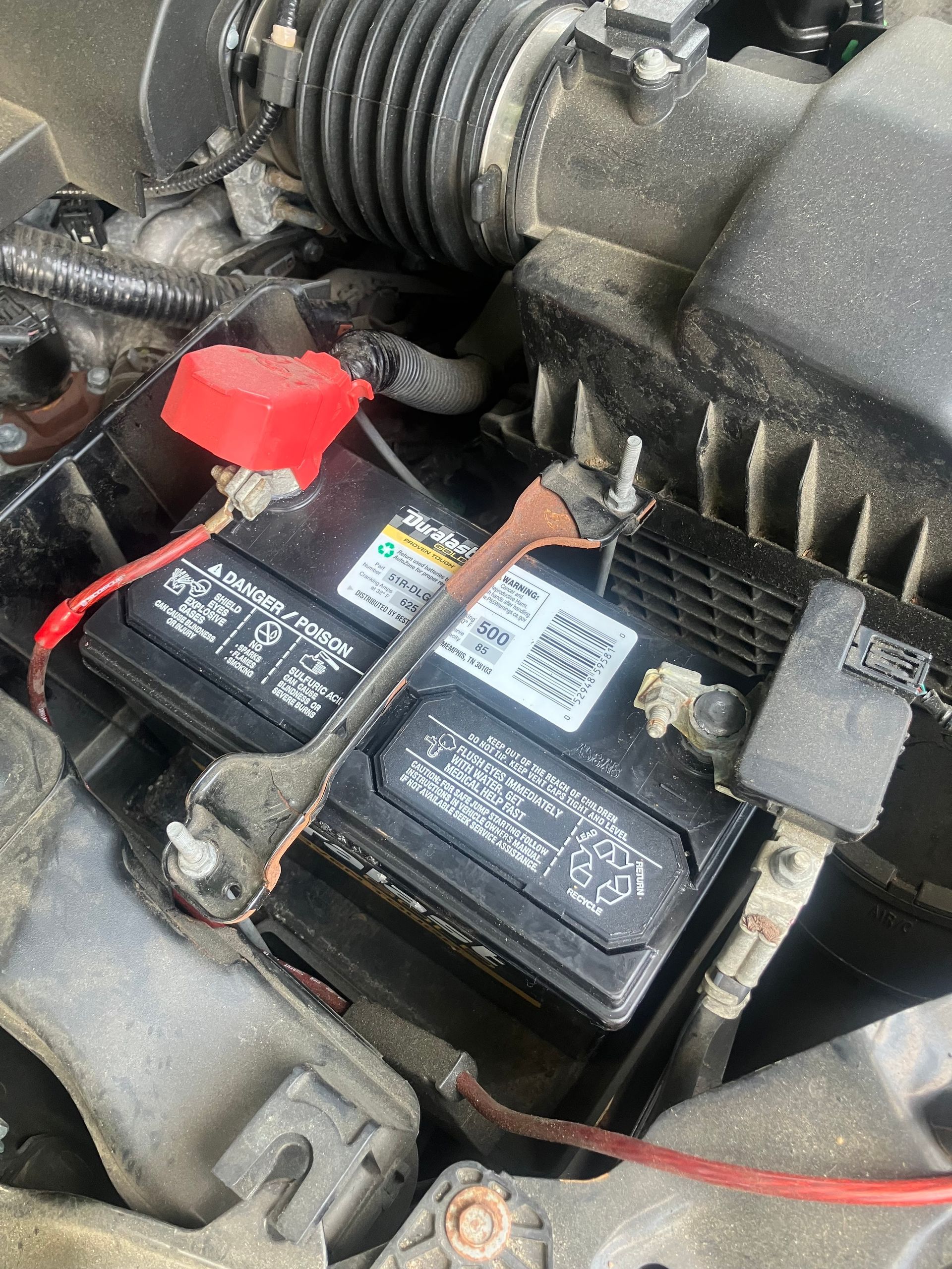 Car engine bay with battery, red positive terminal cover, and connected hoses and wires