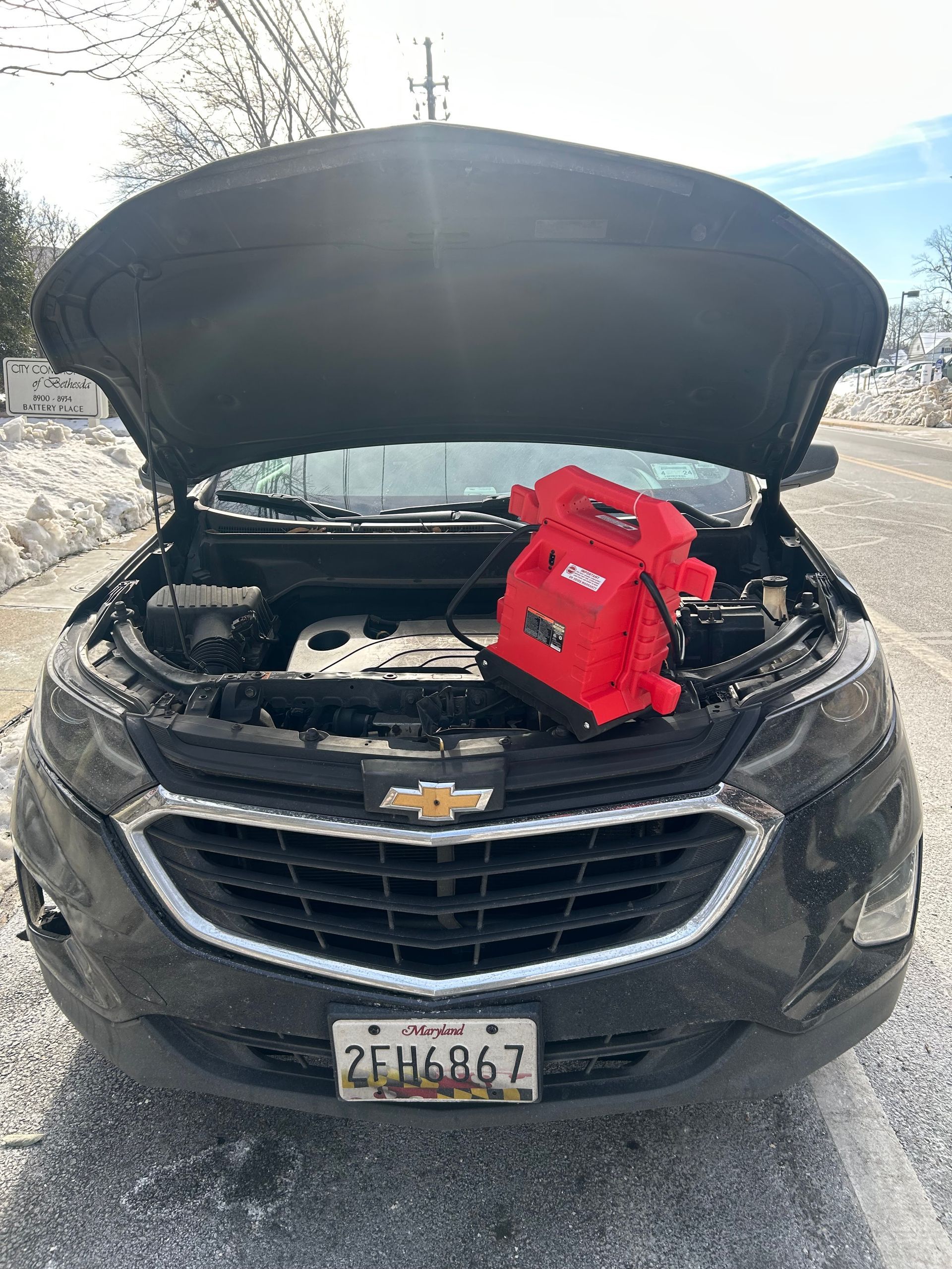 Black Chevrolet SUV with hood open and a red battery booster pack in the engine bay on a snowy roadside