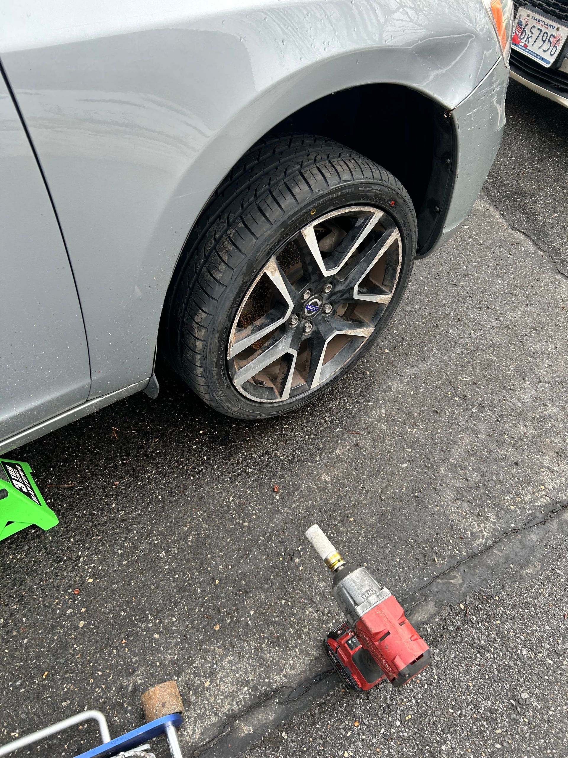 Silver car with flat front tire; tools and a green jack on wet pavement nearby