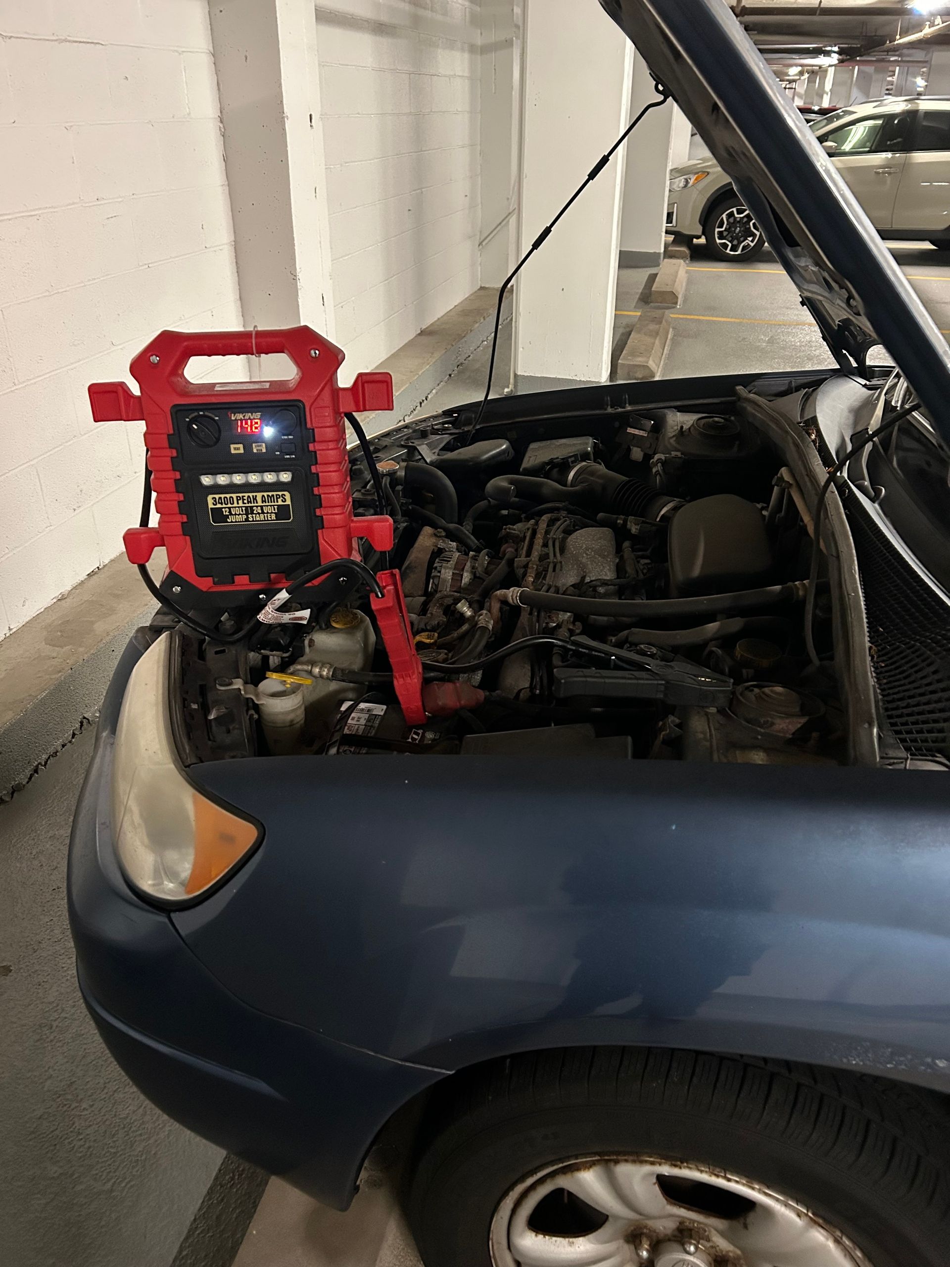 Blue car in garage with hood open and red jump starter attached to the battery.