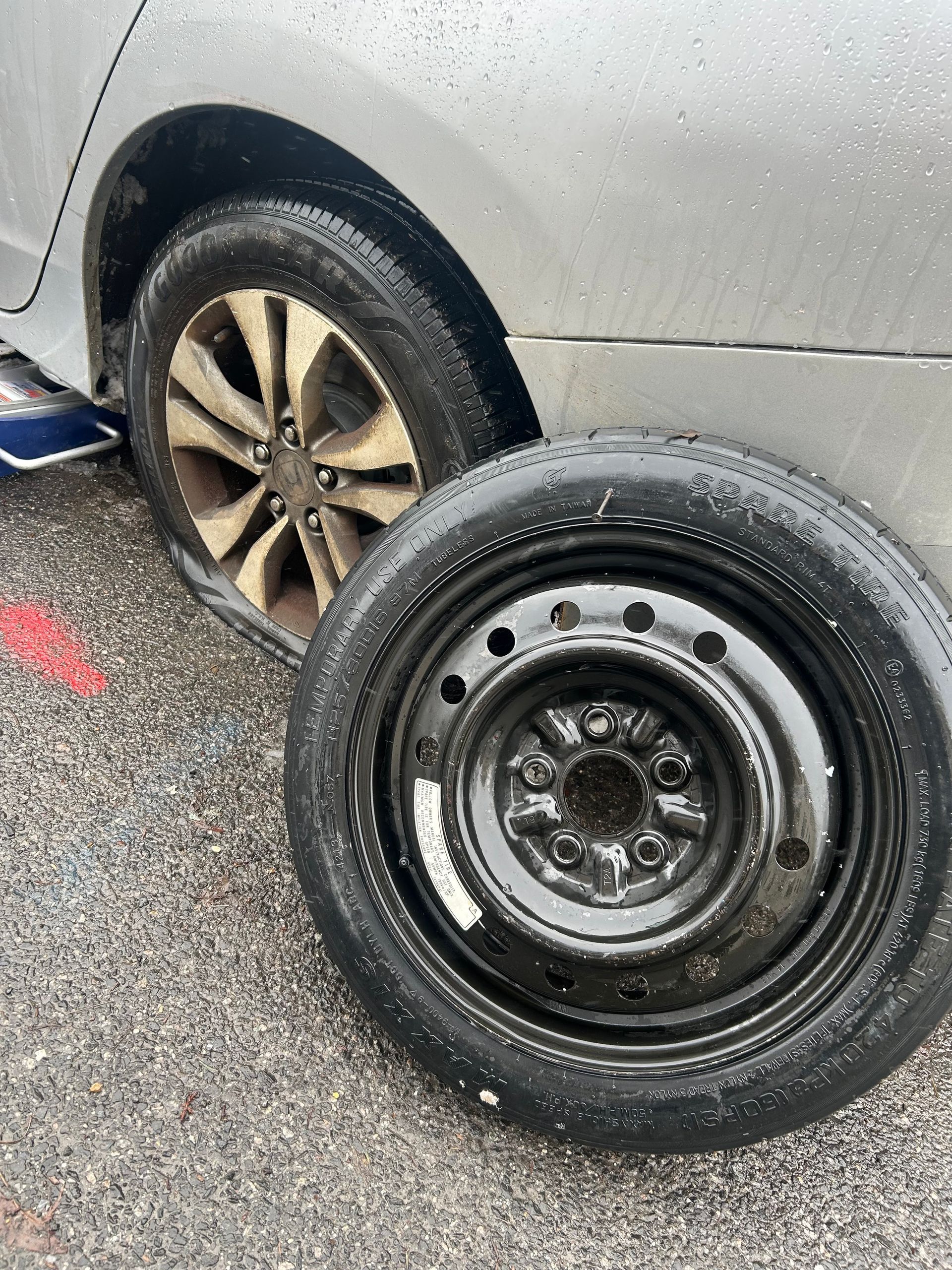 Flat tire beside a silver car on a wet roadside, with the removed wheel leaning in front.