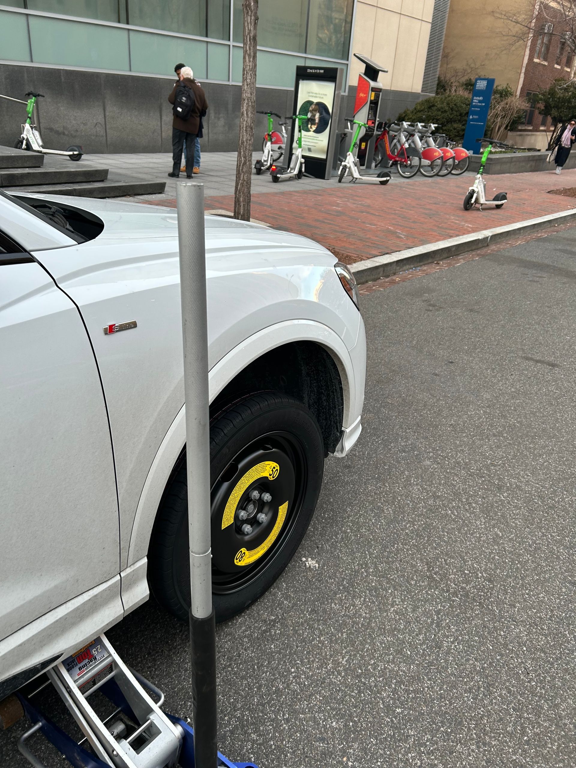 White car with a yellow-rimmed front wheel parked on a city street near a building and people in the background