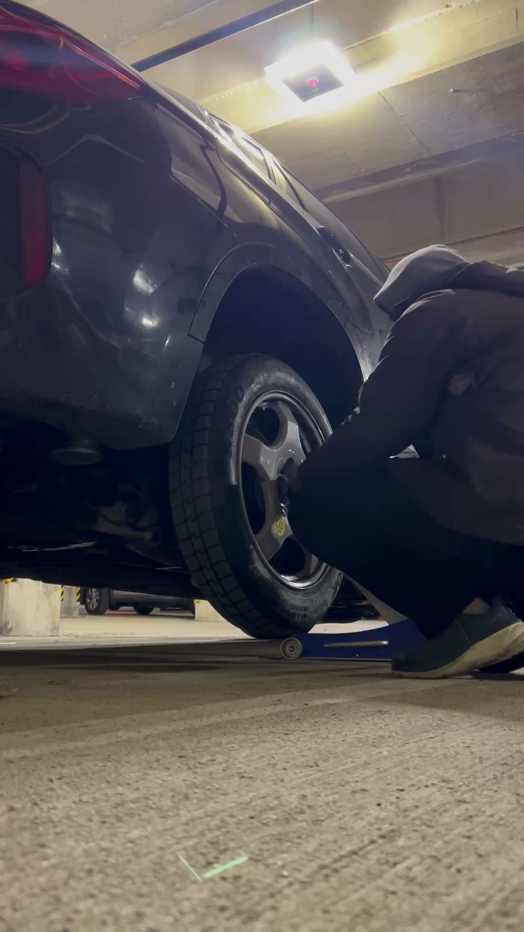 Person crouching beside a car, inspecting or working on the rear wheel in a garage.