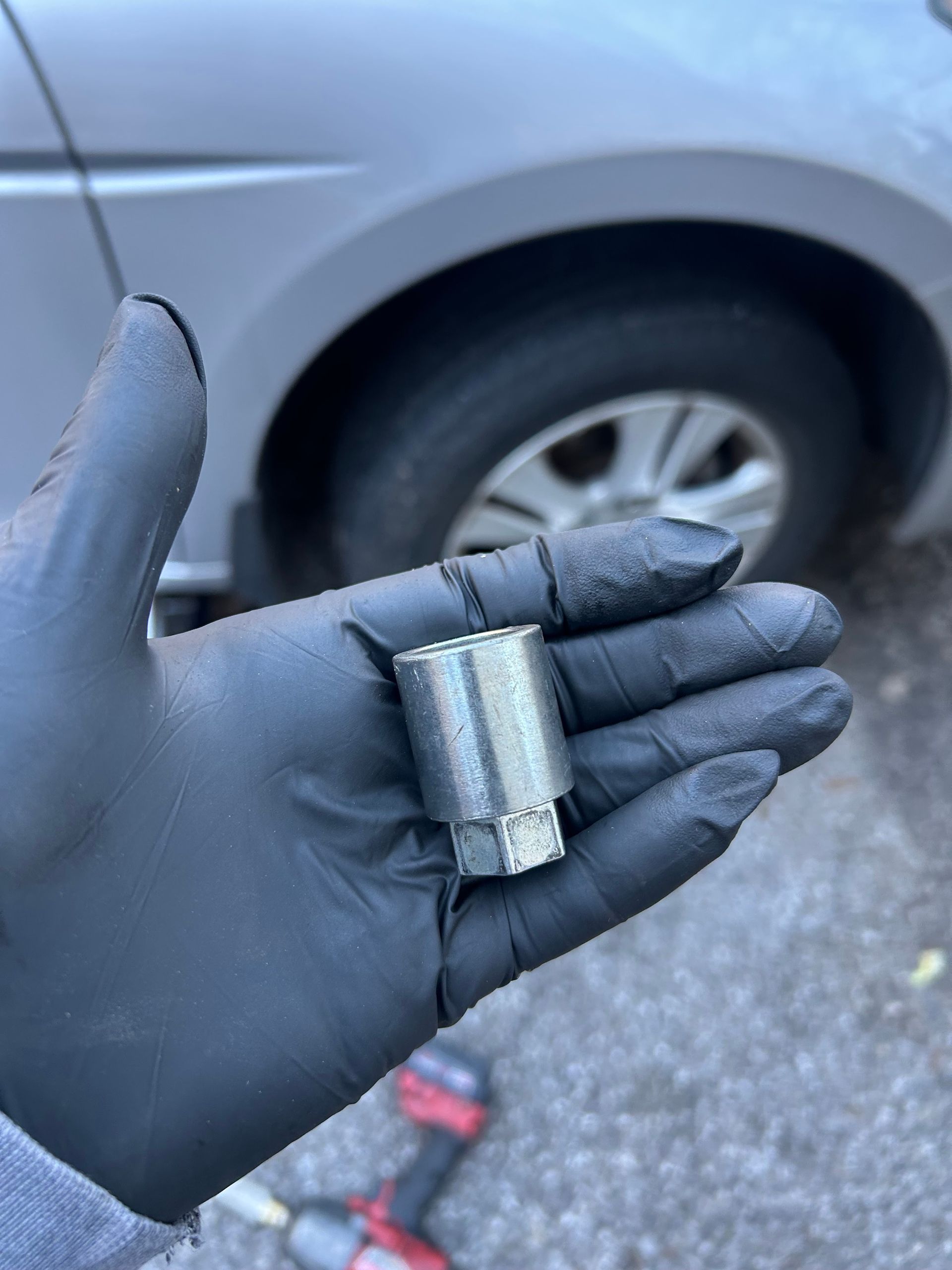 Gloved hand holding a small metal automotive part in front of a car wheel.