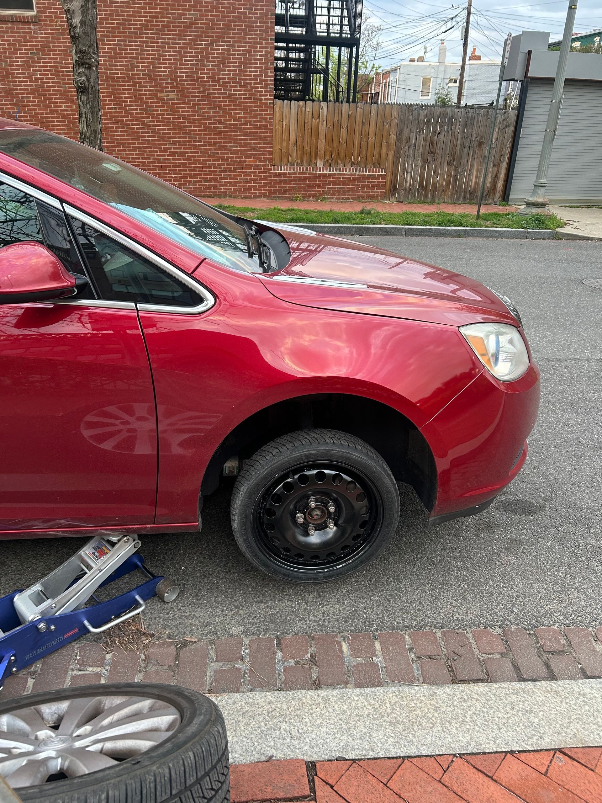 Red car parked by a curb beside a brick building, with a visible spare wheel on the front axle.