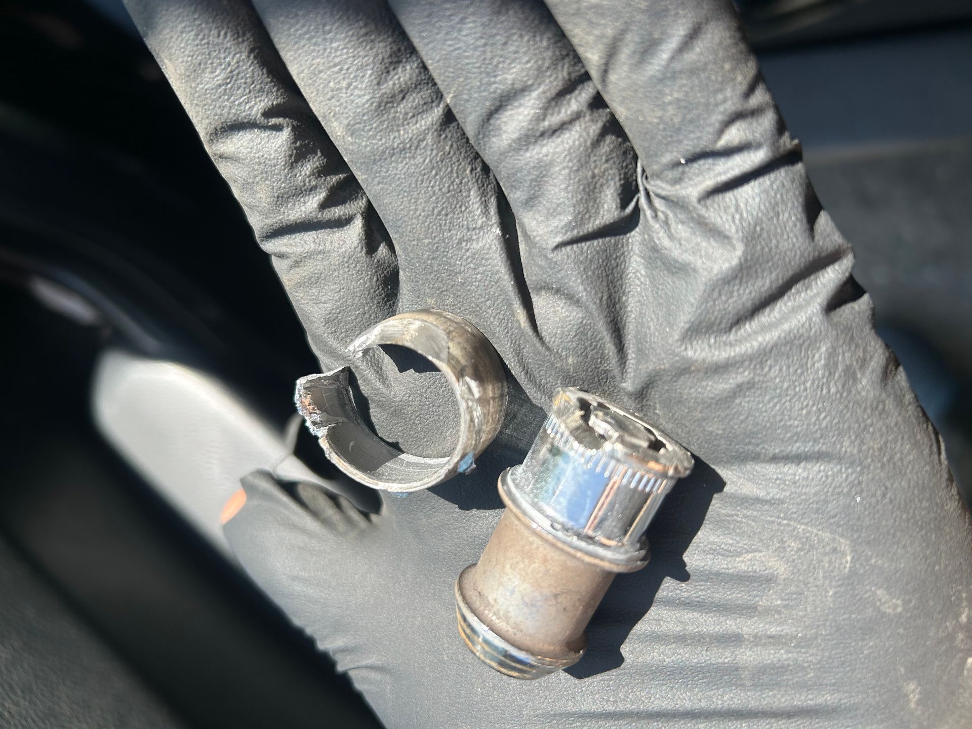 Gloved hand holding two small broken metal parts with a blue ring, likely automotive components.
