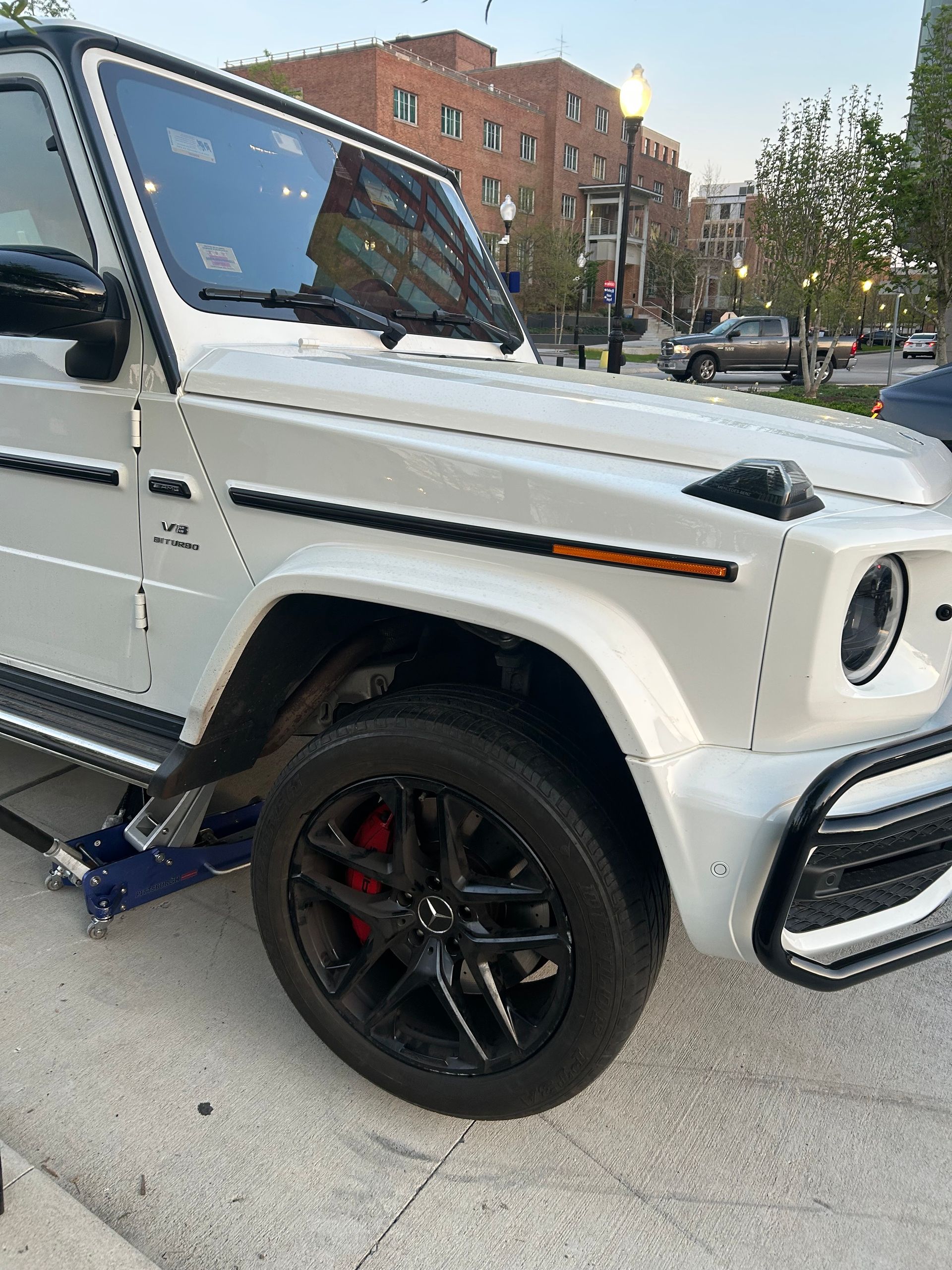 White Mercedes G-Class parked on a city street, front wheel lifted on a blue jack.
