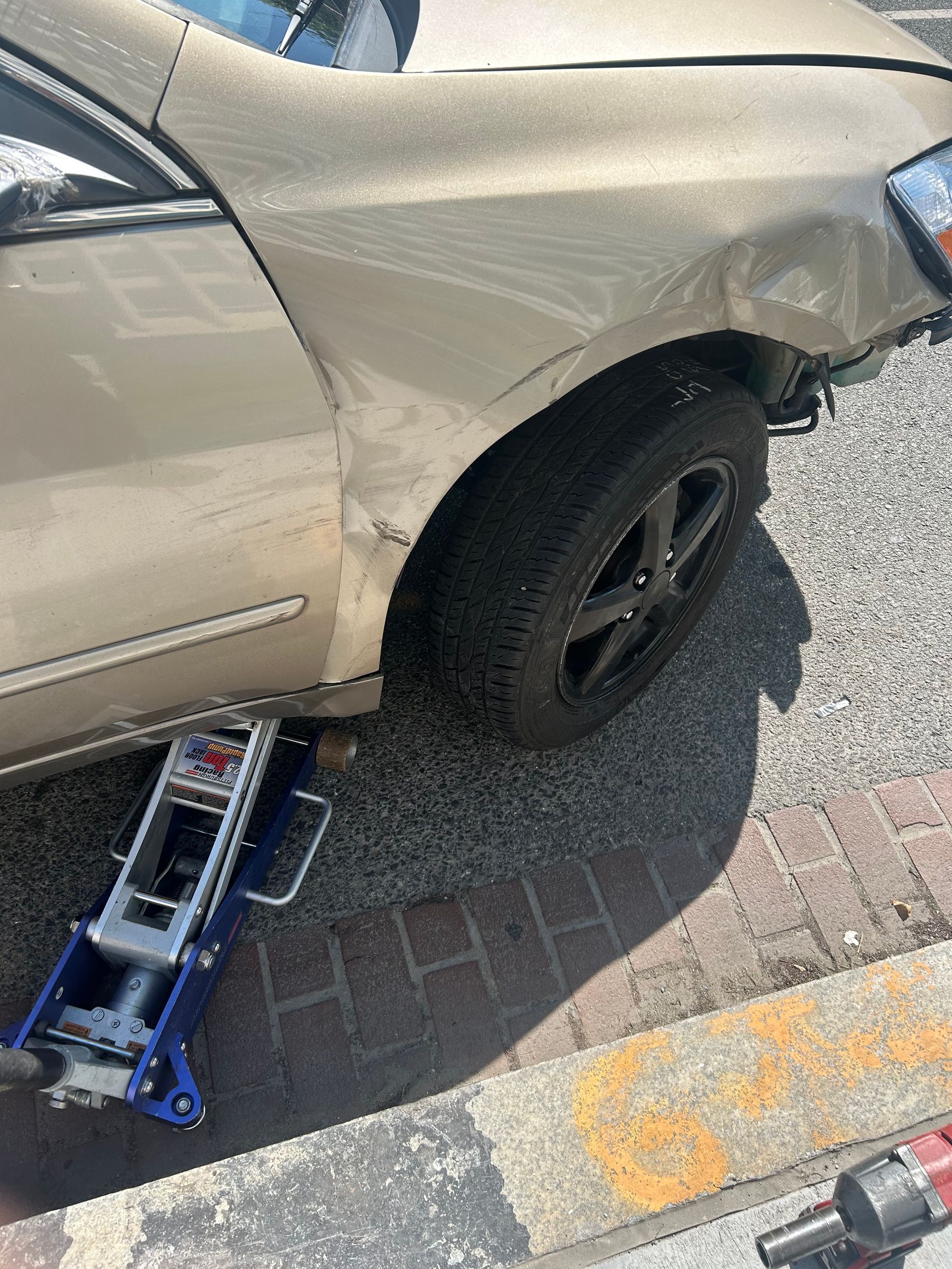 Beige car with a flat rear tire, a floor jack under the side, parked by a curb.