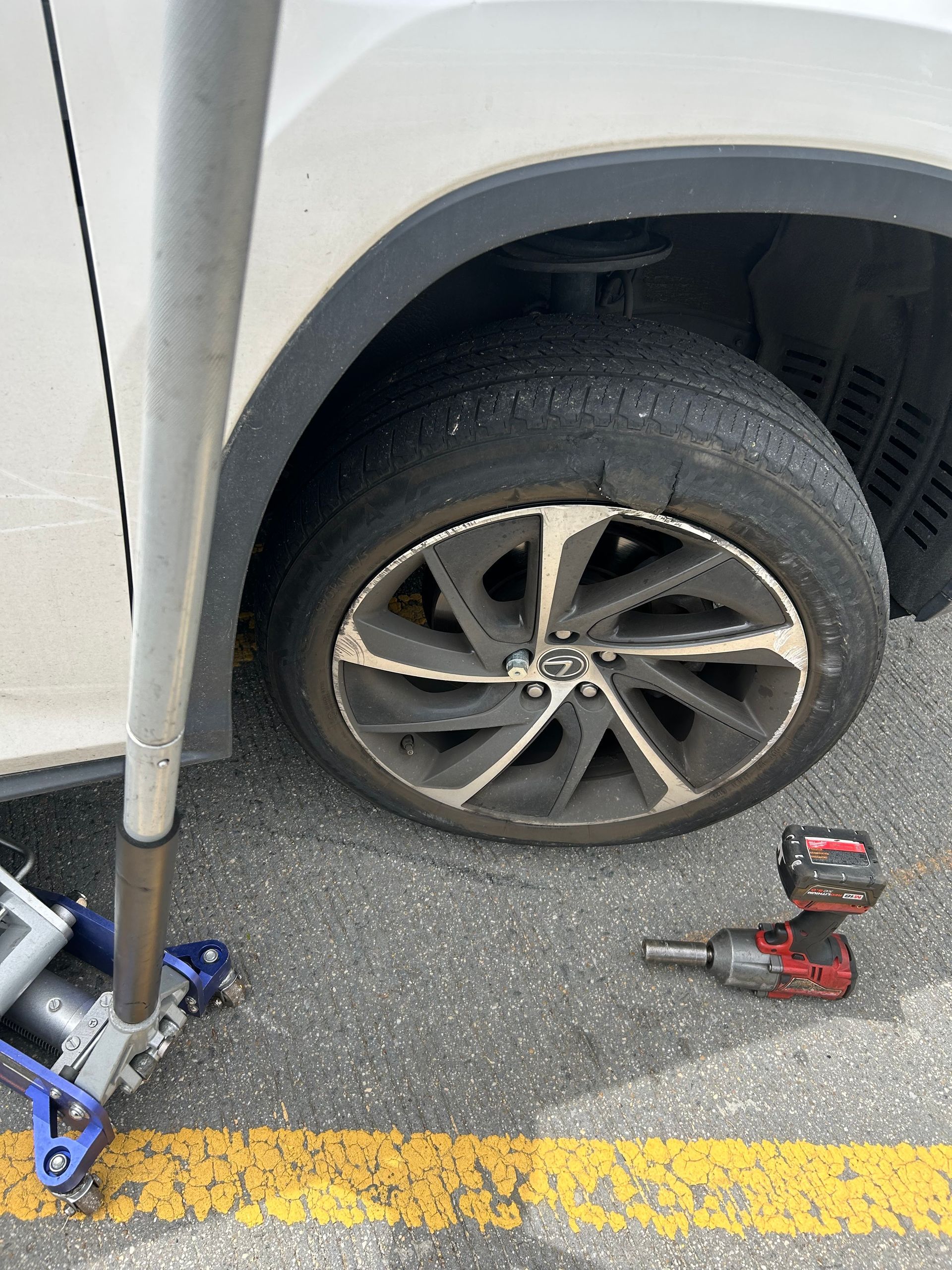 Flat rear tire on a white car beside a yellow line, with a jack and impact wrench on the ground.