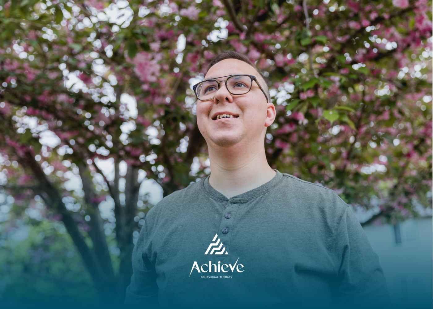 A person wearing glasses and a green shirt smiles while looking up, standing in front of a tree.