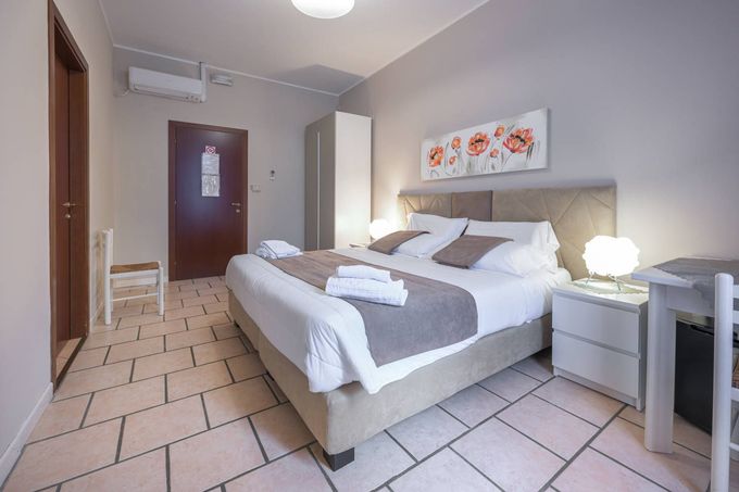 Una camera d'albergo ordinata con un letto ben rifatto, pavimento piastrellato, lampade da comodino, un armadio e un quadro alla parete.