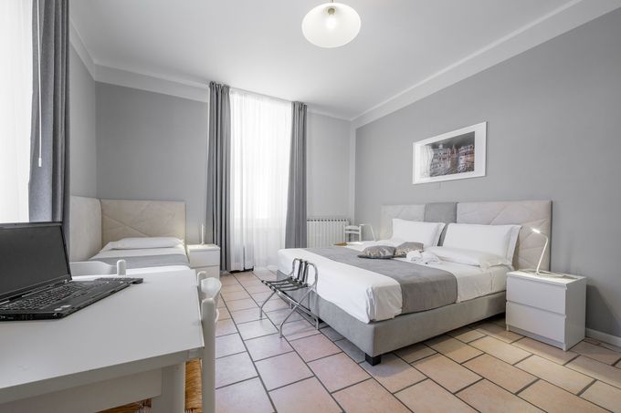 Una camera d'albergo moderna, dai toni neutri, con un letto matrimoniale queen-size, un letto singolo, un computer portatile su una scrivania bianca e pavimento piastrellato chiaro.