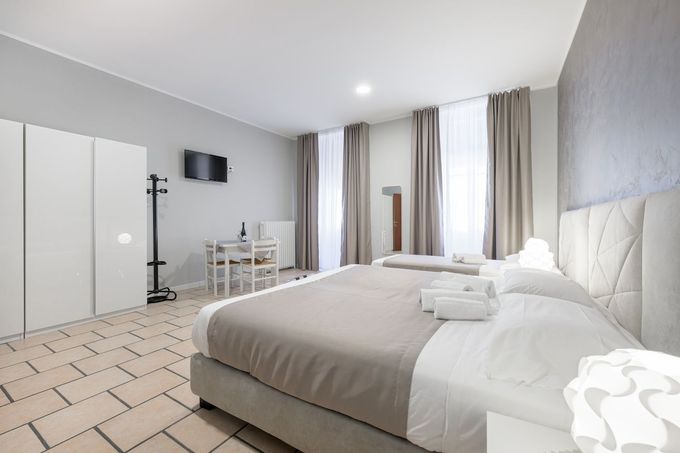 Una camera d'albergo con un grande letto, un armadio bianco, un tavolo da pranzo e due finestre con tende beige su un pavimento piastrellato.