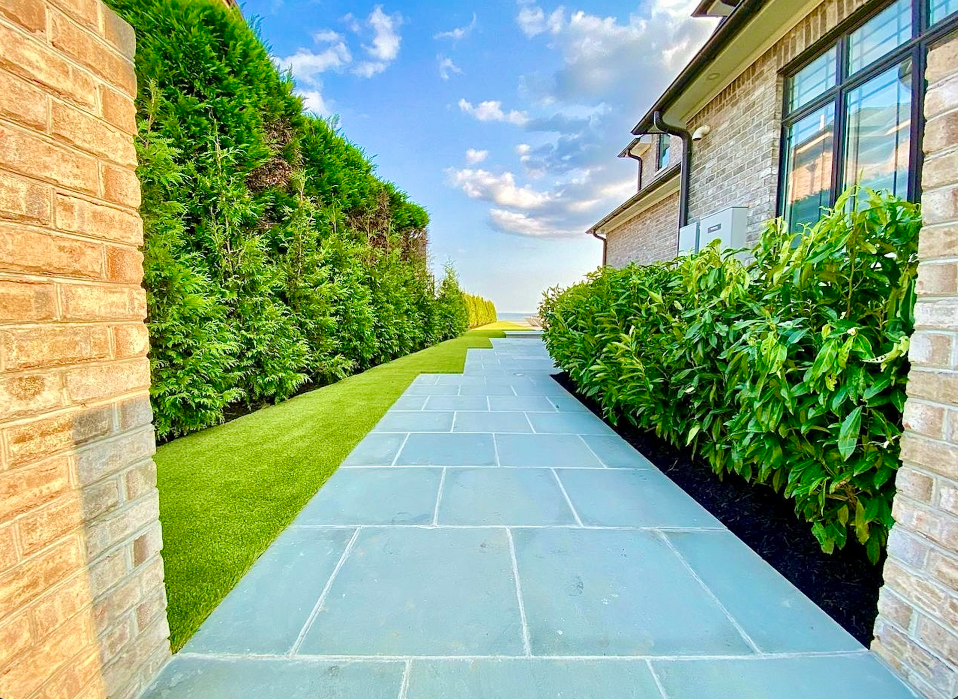 Curved stone walkway through a landscaped garden with shrubs, flowers, and trees beside a house