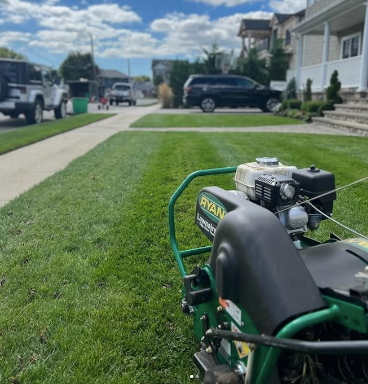 Small gas-powered machine on a green lawn beside a suburban sidewalk and street.