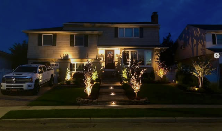 A lit suburban house at night with illuminated front steps, landscaping, and parked vehicles.