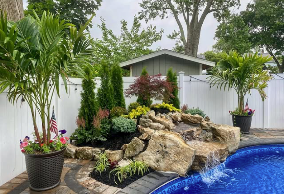 Backyard pool with rock waterfall, tropical plants, and a gray shed behind a white fence