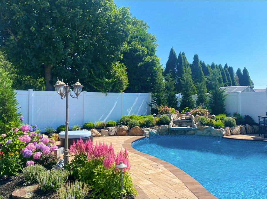 Backyard pool with blue water, pink flowers, white fence, and lush green trees under a clear sky