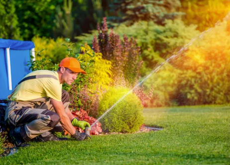 Person kneeling on a lawn, watering plants with a sprinkler in a sunny garden
