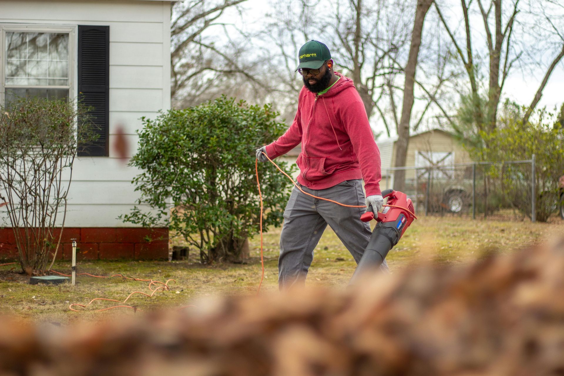 Adult in red sweatshirt guiding a child in a yard beside a white house, with fallen leaves on the ground